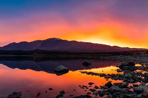 This October 2021 image shows the sunrise powering through rain clouds in Lake Tekapo, Aotearoa New Zealand. The surface of the lake is mostly calm, broken only by raindrops in the middle of the image. A bit of snow remains on Mount Edward in the distance. The reds, oranges, and yellows of this image were only visible for a few minutes because of the shifting rain clouds.