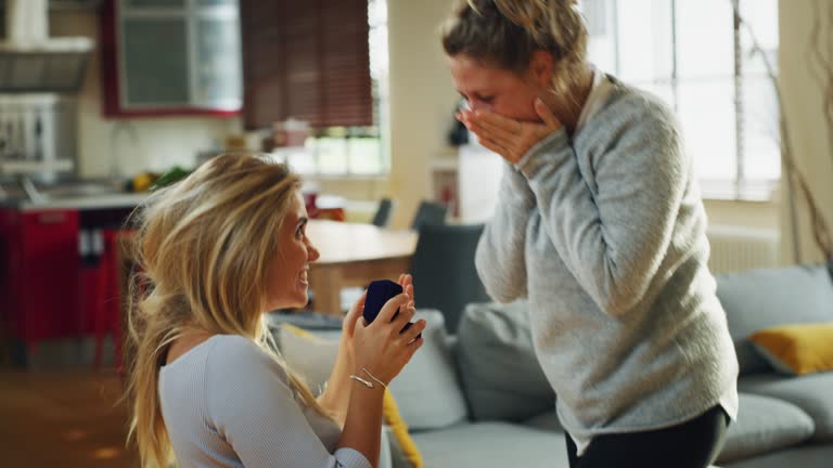 Authentic shot of young homosexual female gay is making a surprise proposal of marriage to her beloved woman in living room at home. The woman is accepting emotionally and giving a hug full of love.