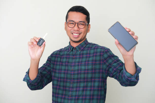 hombre asiático adulto sonriendo feliz mientras muestra la pantalla del teléfono móvil en blanco y sostiene la llave de un automóvil - mid adult men smart casual white happiness fotografías e imágenes de stock