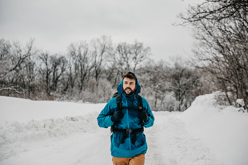 Spontaneous image of a handsome bearded young adult male, wearing a cool winter ski active wear, a yellow kaki pants and a blue jacket, hiking and enjoying the snow outdoors on a mountain, having a backpack with him. Looking at the friend, going a bit slower, while enjoying the sight