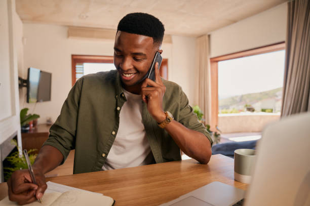 felice giovane maschio nero adulto sorridente durante una telefonata. scrivere nel blocco note, lavorare dal laptop a casa in un appartamento moderno - ewan foto e immagini stock