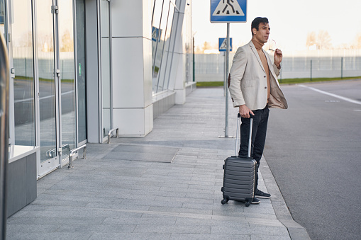 Caucasian male staying alone in open air next to building with grey suitcase while removing glasses from face
