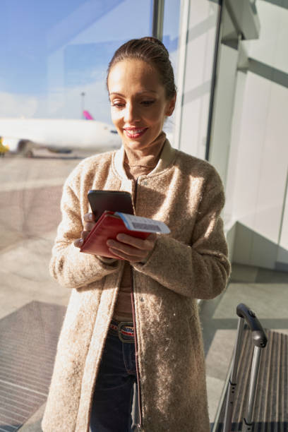 istock Cheerful lady in coat using smartphone near airport window 1365084294
