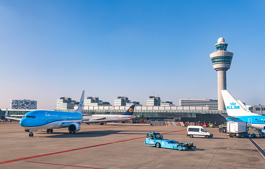 KLM Airplanes at Amsterdam Schiphol airport in Holland. The planes are parked at the gates and a Boeing 737 is taxxiing towards the runways around the terminal building with the air traffic control tower in the background.