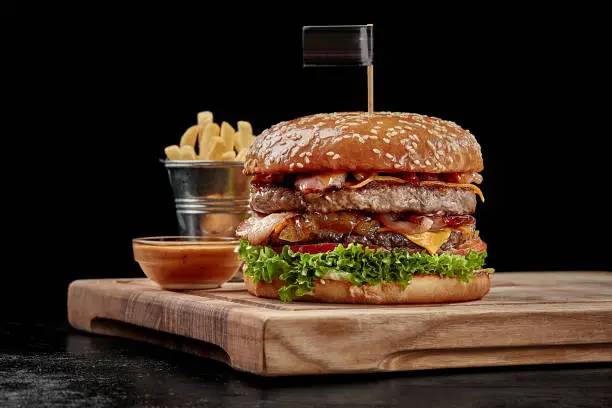 Delicious burger with two juicy pork and beef patties, fried bacon, cheese slices, caramelized onions, fresh tomatoes and greens in fluffy sesame bun served with fries and aioli sauce on wooden board