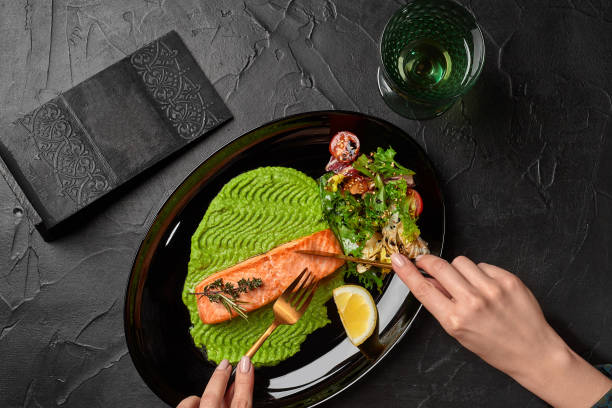 Top view of female hands cutting grilled salmon steak served with mashed peas, salad and lemon stock photo