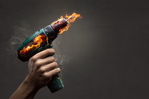 A hand holding a cordless screwdriver that is burning with actual flames. Isolated on a dark background.
