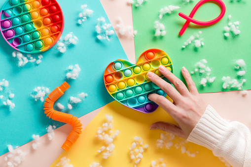 Women's hands press on a silicone rainbow antistress toy in the shape of a heart and other toys on a cardboard of different colors and fluff. Relaxation and tranquility