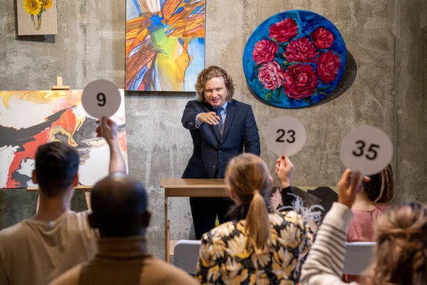 Male auctioneer pointing at one of people with auction paddles Mature male auctioneer in formalwear standing by tribune and pointing at one of people with auction paddles auction stock pictures, royalty-free photos & images