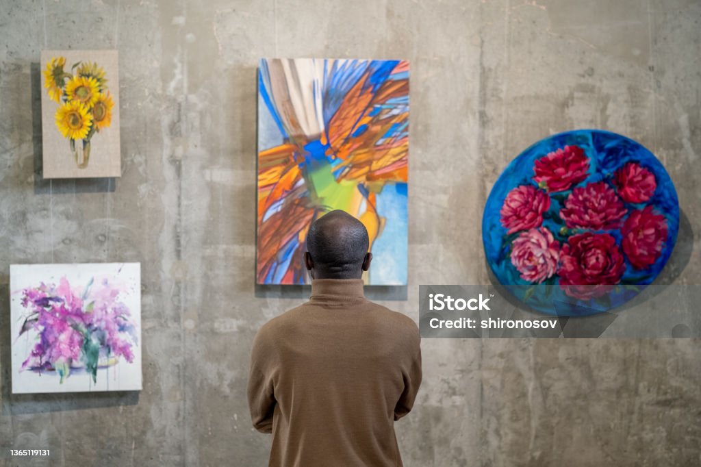 Back view of African male guest of art gallery standing in front of wall with expositions Back view of African male guest of art gallery standing in front of wall with expositions and looking at one of them Art Museum Stock Photo