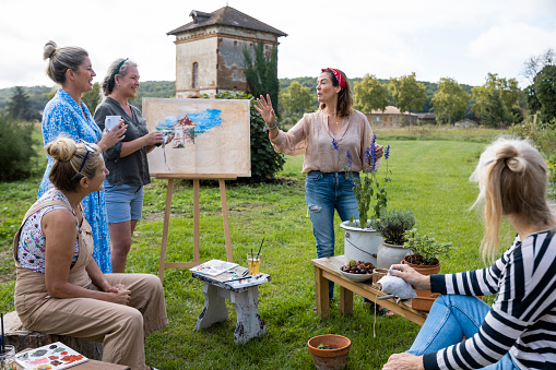 A group of mature women standing outdoors during an art class on a wellness retreat in Castelferrus, France. They are all looking at the art tutor who is teaching the class and explaining tips to them.