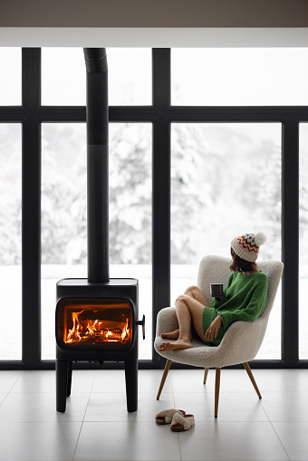Woman sitting with cup on chair by the fireplace at modern house on nature during winter time. Concept of winter mood and comfort at home. Girl wearing hat and green sweater