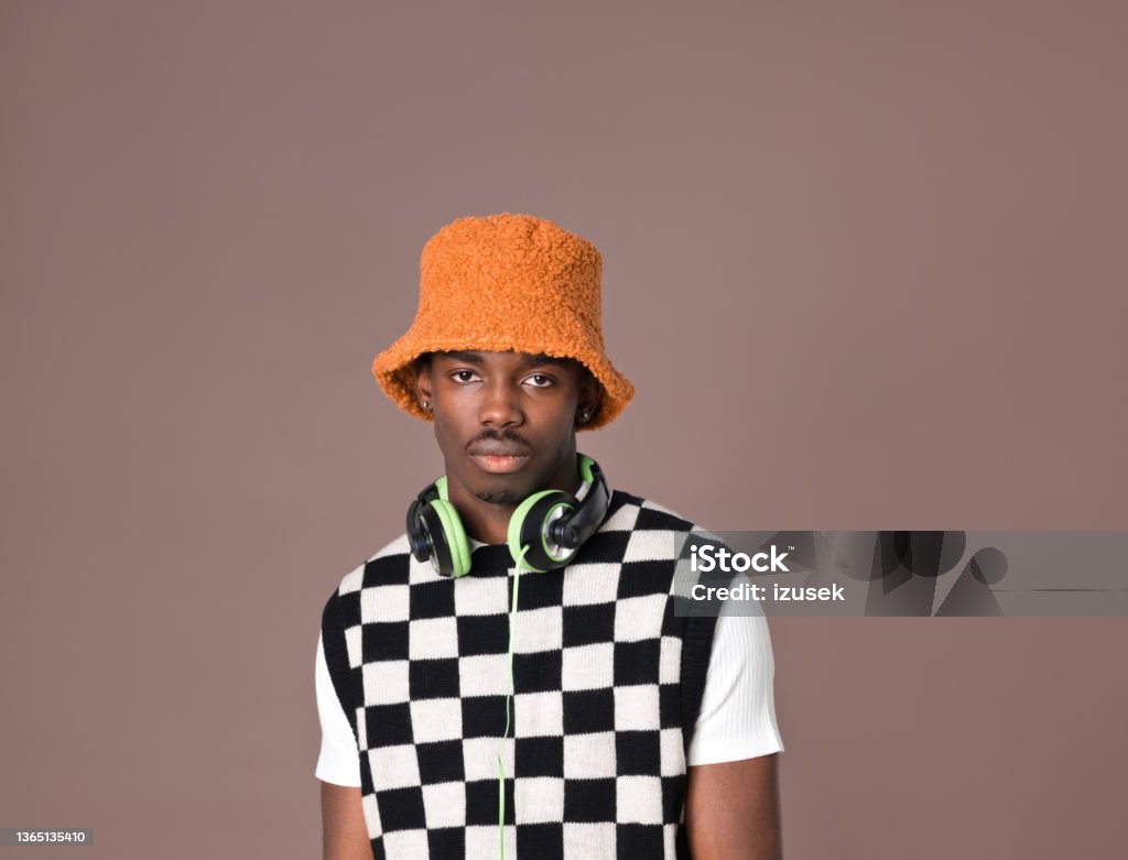 Confident young man against brown background Portrait of young man wearing black and white checker chess sweater vest, orange bucket hat and green headphone looking at camera. Studio shot on brown background. Bucket Hat Stock Photo
