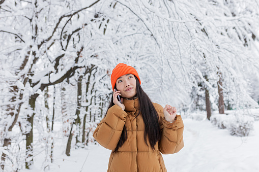 Portrait of a young beautiful Asian woman, photo of a close up having fun talking on the phone in a winter snow-covered park warmly dressed