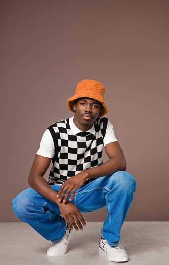 Portrait of fashionable young man wearing checker chess sweater vest blue jacket, jeans pants  and orange bucket hat looking at camera. Studio shot on brown background.