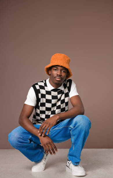 Confident young man against brown background Portrait of fashionable young man wearing checker chess sweater vest blue jacket, jeans pants  and orange bucket hat looking at camera. Studio shot on brown background. bucket hat stock pictures, royalty-free photos & images