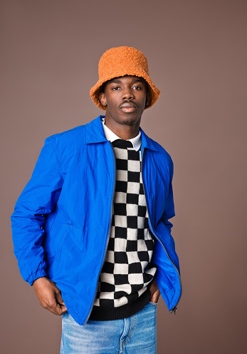 Portrait of fashionable young man wearing checker chess knit sweater, blue jacket and orange bucket hat looking at camera. Studio shot on brown background.