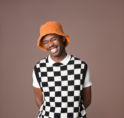 Portrait of happy young man wearing checker chess sweater vest and orange bucket hat laughing with eyes closed. Studio shot on brown background.