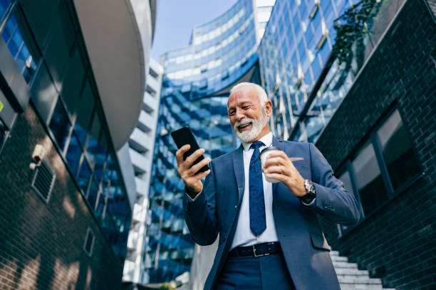 An old executive reading message on the phone at the business center. Low angle view of a happy senior businessman standing at the business center and reading a message on the phone. stock photo