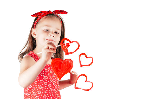 Little girl with long curly hair in red clothes draw hearts with paint on transparent glass smiling on white background. stock photo