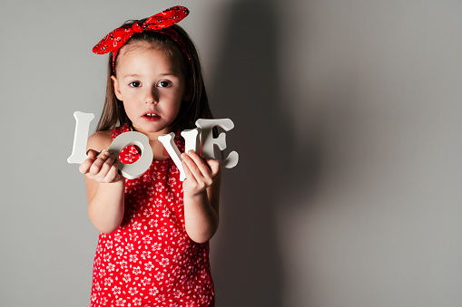 Pretty little girl with long hair in red clothes hold white volumetric letters of word love in hands on gray background with shadow. Family, love, holidays, birthday, Mother's day concept. Copy space.