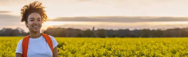 Panorama header beautiful mixed race African American girl teenager female young woman smiling and happy hiking with back pack in a field of yellow flowers at sunset or sunrise panoramic web banner.