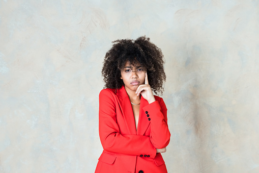 Fashion portrait of angry curly hair young woman wearing red suit, standing with hand on chin and looking at camera. Studio shot on beige background.