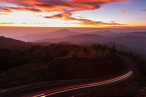 The scenery of mountain road at dawn, curve mountain road with lights trails, dramatic clouds in the sunrise sky in the background. Transportation, travel concepts. Long exposure.