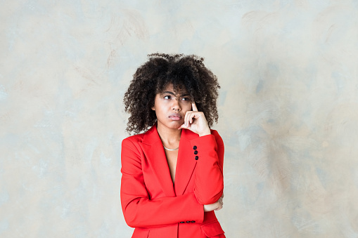 Portrait of displeased curly hair young woman wearing red suit, standing with hand on chin and looking away. Studio shot on beige background.