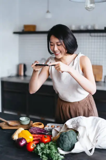 Young Asian woman coming home from grocery shopping, taking out fresh fruits and vegetables from a reusable shopping bag on the kitchen counter. She is taking photos of the fresh produces with smartphone at home