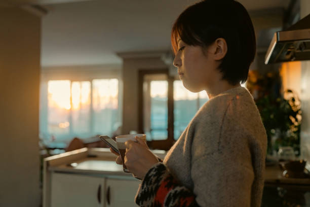 istock Beautiful young woman using smart phone and relaxing in kitchen during sunset 1365169560
