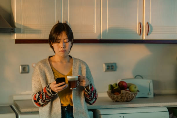 istock Beautiful young woman using smart phone and relaxing in kitchen during sunset 1365169719