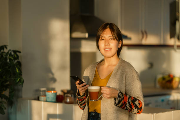 istock Beautiful young woman using smart phone and relaxing in kitchen during sunset 1365170672