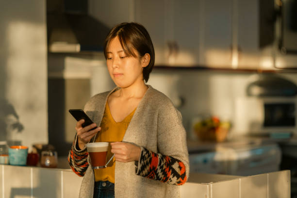 istock Beautiful young woman using smart phone and relaxing in kitchen during sunset 1365170754