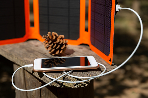 Smartphone is charging from solar battery in summer camp. Clean energy for using in camping. Gadgets are lying on the old wooden bench..