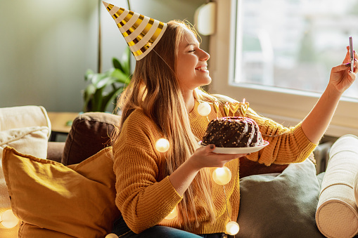 A young woman is at home in the living room, she is holding a birthday cake and taking a selfie