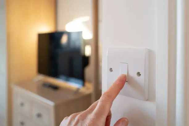 Shallow focus of a home owner switching off a bedroom light after waking up in late morning. A smart TV can be seen in the room.