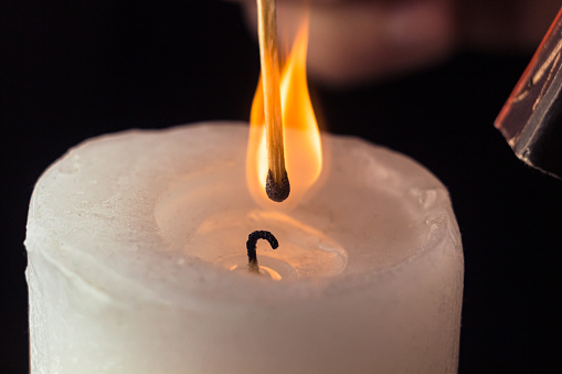 Hand holding burning match stick lighting candle on black background. Match with the flame and candle in dark room close up. Ignition of a match