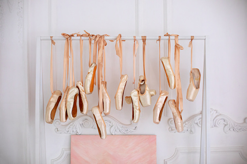 New pointe shoes with satin ribbons hanging on rank. Ballet shoes hang on bar in room. Concept of dance, ballet school, ballerinas clothes. Many hanging ballet shoes on white wall background in studio