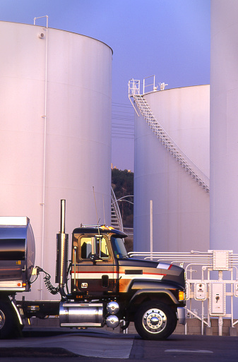 A fuel tanker truck is parked next to several white fuel storage tanks.