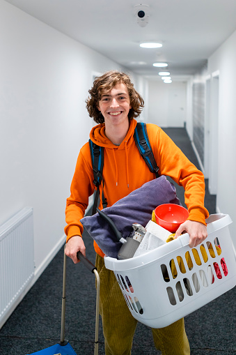 A medium close up portrait of a young male with wavy hair as he moves out from home for the first time. He is carrying some of his belongings in a laundry basket which will be used in his room.