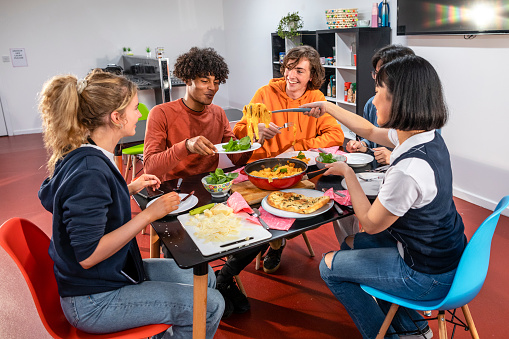 An over-the-shoulder view of a group of young university students getting together for a meal in their student accommodation in Sunderland in the North East of England. One of the female students is dishing up the pasta to everyone one by one.