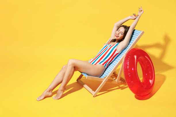 Full body length happy young sexy woman wear striped red blue one-piece swimsuit sit on wooden chair isolated on vivid yellow color wall background studio. Summer hotel pool sea rest sun tan concept. Full body length happy young sexy woman wear striped red blue one-piece swimsuit sit on wooden chair isolated on vivid yellow color wall background studio. Summer hotel pool sea rest sun tan concept chaise longue woman stock pictures, royalty-free photos & images