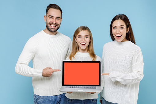 Excited young parents mom dad with child kid daughter teen girl in sweaters point index fingers on laptop computer with blank empty screen isolated on blue background. Family day parenthood concept