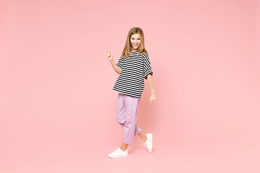 Full length little blonde kid smiling cute girl 12-13 years old in striped oversized t-shirt walking going clench fist isolated on pastel pink background children portrait. Childhood lifestyle concept