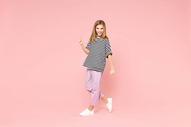 full length little blonde kid smiling cute girl 12-13 years old in striped oversized t-shirt walk going clench fist isolated on pastel pink background children portrait. childhood lifestyle concept - teenager 14 15 years 13 14 years cheerful imagens e fotografias de stock