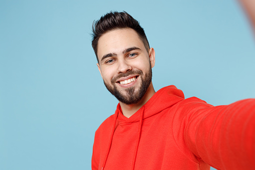 Close up young caucasian smiling happy bearded attractive student handsome man 20s wearing casual red orange hoodie looking camera isolated on blue background studio portrait People lifestyle concept