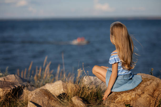 Little beautiful girl with long blond hair sits on the seashore and looks at the waves