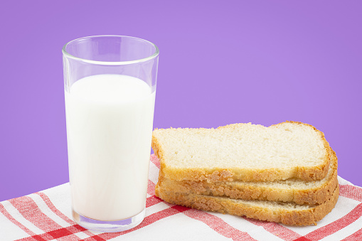 Full glass of milk with slices of white rustic bread on red-white striped linen napkin on purple background and copy space