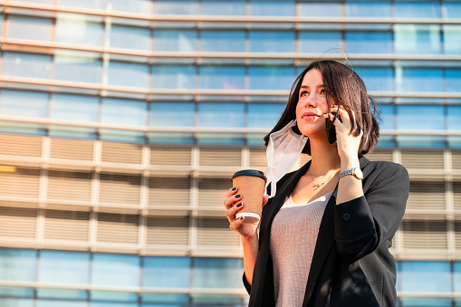 Portrait of pretty young businesswoman remove medical protective mask down to infection prevention talking with a smart phone, holding a coffee cup and working in Covid-19 outbreak after novel coronavirus quarantine